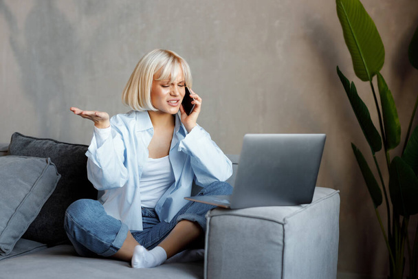 Belle jeune fille blonde parlant au téléphone et travaillant sur un ordinateur portable à la maison sur le canapé. pigiste, travail, style de vie moderne. - Photo, image