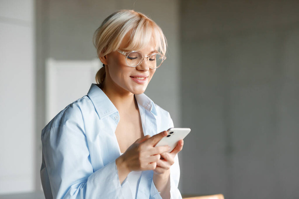 Beautiful young blond girl in stylish glasses is working on her phone. Smiling and looking at the phone. - Foto, imagen