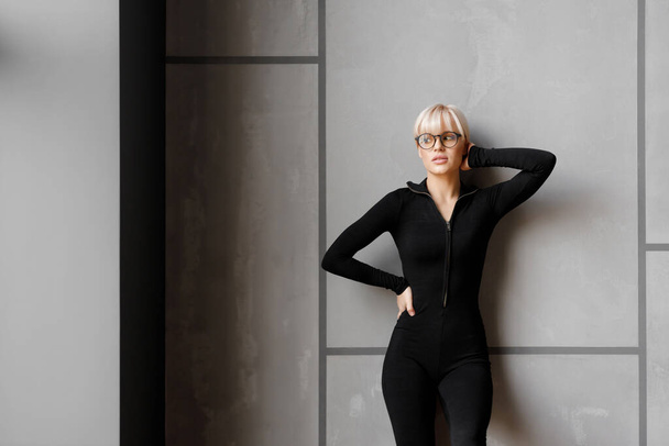 A beautiful young blond girl in stylish glasses and a tight black suit looks away. Fashion photography concept. - Valokuva, kuva