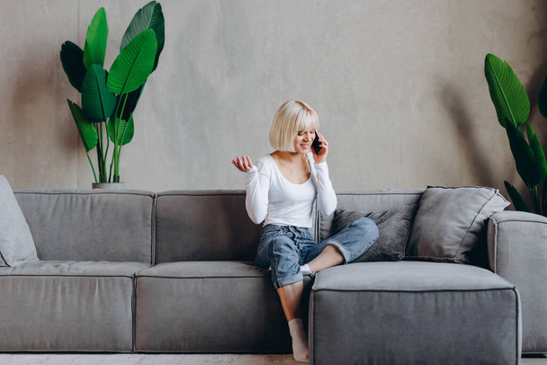 Beautiful blonde girl is sitting on the sofa and talking on the phone. Freelance concept. - Фото, изображение