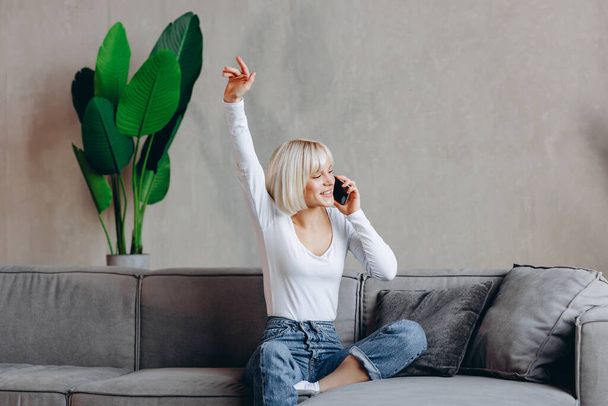 Красивая блондинка счастливая девушка сидит на диване и разговаривает по телефону. - Фото, изображение