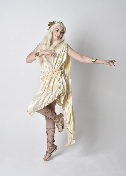 Full length πορτρέτο της όμορφης ξανθιάς γυναίκας φορώντας ένα κοστούμι θεά φαντασία toga με μαγικό στεφάνι.Μόνιμη, χορευτική στάση με χειρονομιακά χέρια που φθάνουν έξω, απομονώνονται σε λευκό φόντο στούντιο. - Φωτογραφία, εικόνα