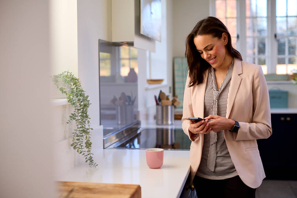 Businesswoman At Home With Looking At Mobile Phone Standing By Kitchen Counter - Photo, Image