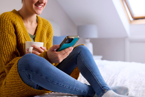 Close Up Of Young Woman Sitting On Bed At Home Smiling Looking At Mobile Phone Holding Hot Drink - Фото, изображение