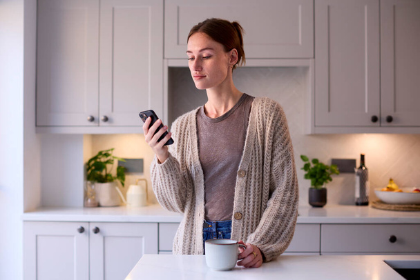Молода жінка розслабляється вдома на кухні тримає гарячий напій за допомогою мобільного телефону
 - Фото, зображення