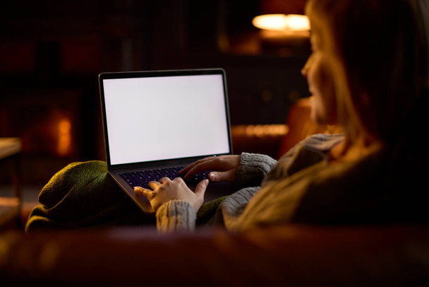 Femme à la maison dans le salon allongé sur le canapé avec le feu confortable regardant l'écran vide sur l'ordinateur portable - Photo, image