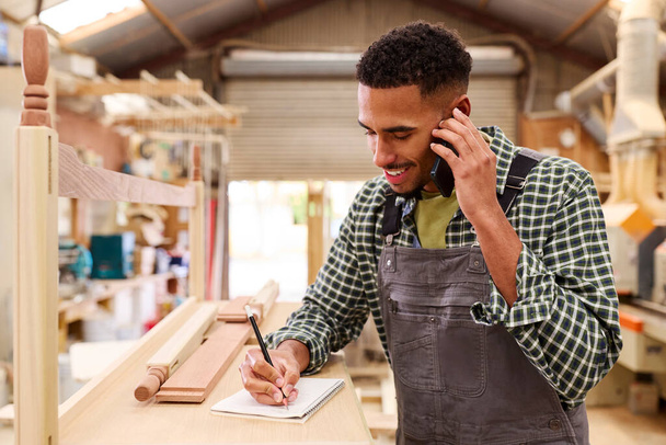 Male Apprentice Working As Carpenter In Furniture Workshop Making Phone Call - Foto, Imagen
