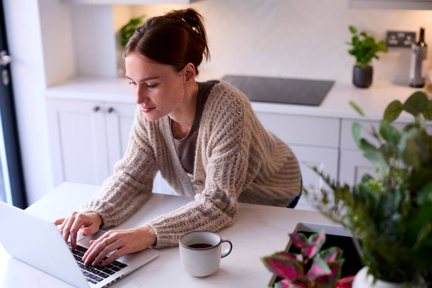 Young Woman At Home Working On Laptop On Counter In Kitchen - Foto, afbeelding