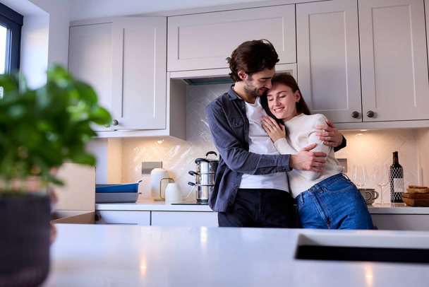 Young Couple Moving Into New Home Hugging As They Unpack Boxes In Kitchen Together - Foto, imagen