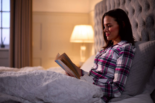Woman At Home Sitting In Bed In Evening Reading Book - Photo, Image