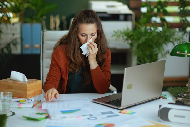 stylish small business owner woman with laptop and napkin blowing nose and working with documents in the modern green office. - 写真・画像