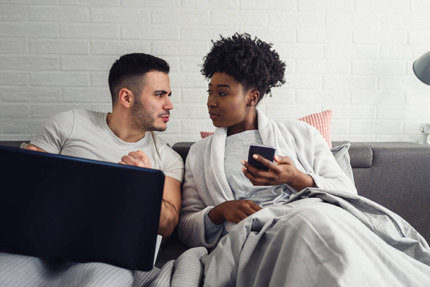 若い多人種のカップルがベッドで彼らのモバイル電子機器を使用しています。インターネット中毒. - 写真・画像