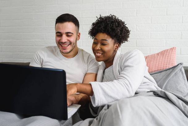 Happy young multiracial couple , lying in a bed and laptop - Foto, imagen