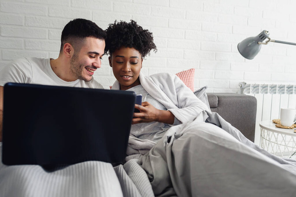 Boldog fiatal multiracionális pár, ágyban és laptopon fekve - Fotó, kép
