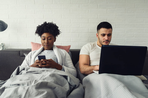 Young multiracial couple using their mobile electronic devices in bed ignoring each other. Relationship and communication problems and internet social network addiction. - Zdjęcie, obraz