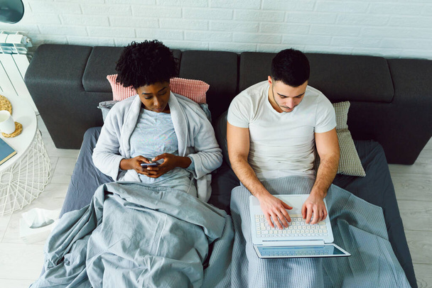 Junge multiethnische Paare, die ihre mobilen elektronischen Geräte im Bett benutzen und sich dabei gegenseitig ignorieren. Beziehungs- und Kommunikationsprobleme und Sucht nach sozialen Netzwerken im Internet. - Foto, Bild