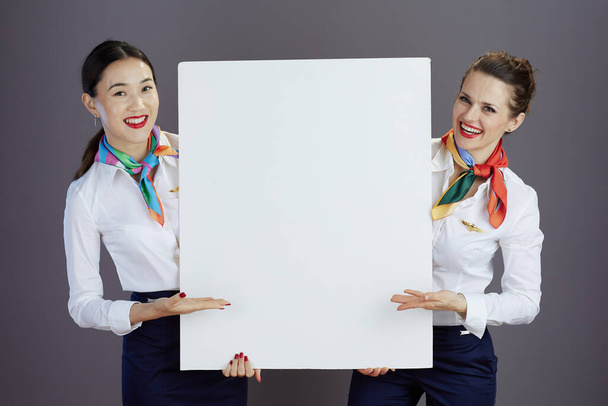 smiling elegant female flight attendants in blue skirt, white shirt and scarf with blank billboard isolated on grey. - Photo, Image