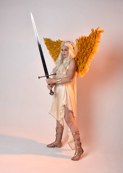 Full length portrait of beautiful blonde woman wearing a fantasy goddess toga costume with feathered angel wings, holding a sword weapon. Jumping pose like flying, isolated on white studio background. - Φωτογραφία, εικόνα