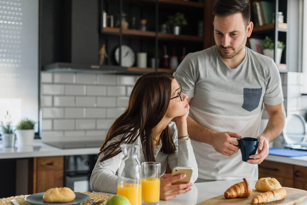 Uśmiechnięta para ciesząca się pięknym porankiem w domowej kuchni, jedząc śniadanie, pijąc kawę i sok i patrząc na smartfona - Zdjęcie, obraz