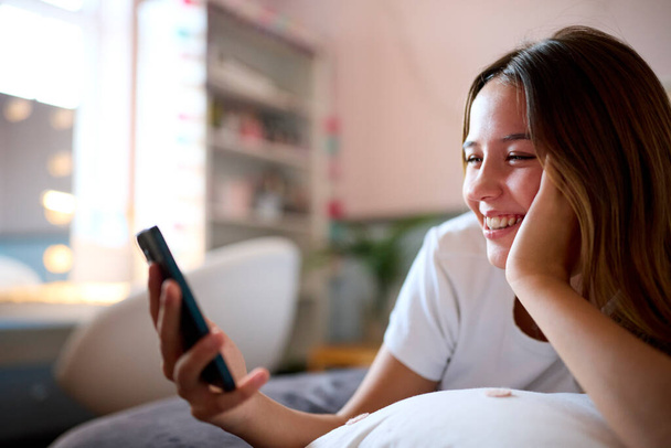 Smiling Teenage Girl Lying On Bed At Home Looking At Mobile Phone - Foto, Bild