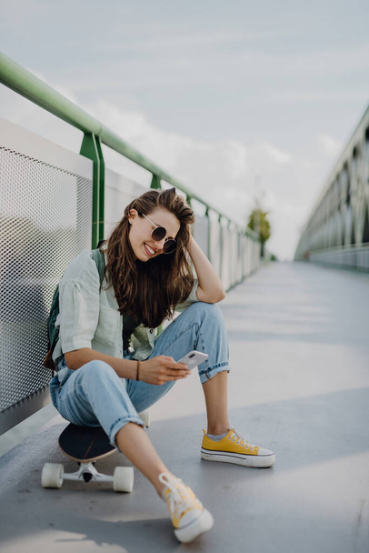 Happy young woman sitting on a skateboard and scrolling smartphone at city bridge. Youth culture and commuting concept. - Zdjęcie, obraz
