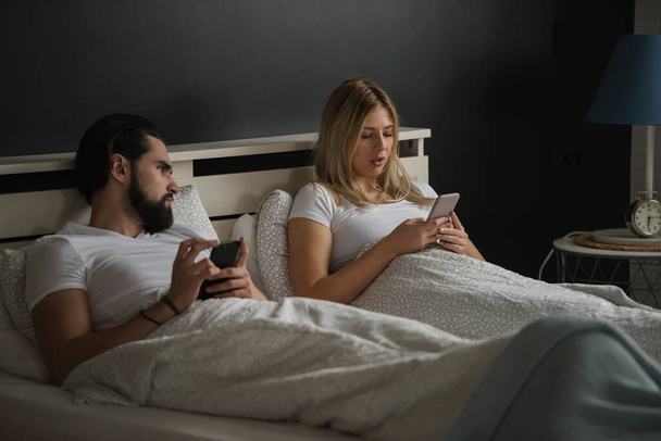若い大人のカップルは、社会的ネットワークに中毒ベッドでスマートフォンを使用しています。モバイル中毒の概念 - 写真・画像