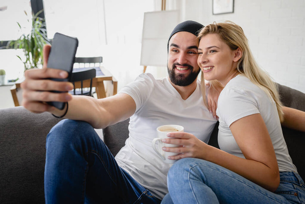 Joven pareja hipster sentada en el sofá en casa tomando una foto con un smartphone - Foto, imagen