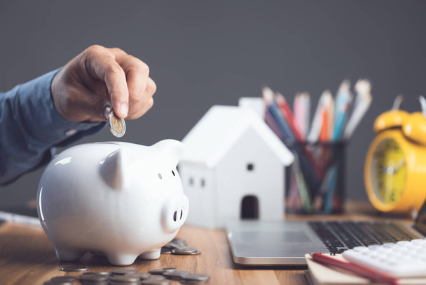 House purchase and money saving idea, Home cost calculate concept. Man adding coin to white piggy bank on office desk to plan for house financial. Residence buying planning in the future. Studio shot. - Zdjęcie, obraz