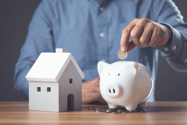 Money saving for house or Financial for real estate concept. Man adding coin to white piggy bank and small house model on the wooden table. Keeping fund for purchase the residence idea. Studio shot. - Photo, image