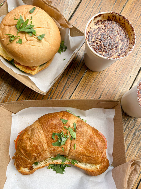 Vista superior del apetitoso sándwich de hamburguesa y croissant en cajas de cartón servidas en mesa de madera con tazas desechables de café espumoso en el restaurante - Foto, imagen