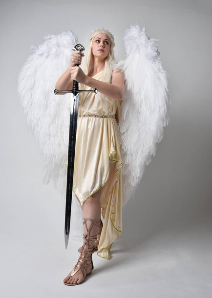 Retrato de cuerpo entero de una hermosa mujer rubia vestida con un disfraz de diosa toga de fantasía con alas de ángel emplumadas, sosteniendo un arma de espada. Saltando posan como volando, aislados sobre fondo blanco del estudio. - Foto, Imagen