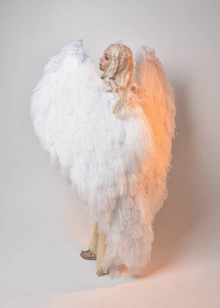 Full length portrait of beautiful blonde woman wearing a fantasy goddess toga costume with feathered angel wings.  Jumping pose like flying, isolated on white studio background. - Photo, Image
