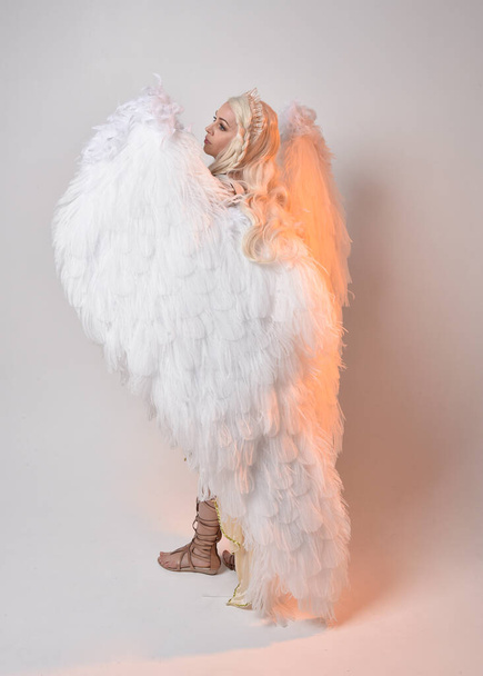 Full length portrait of beautiful blonde woman wearing a fantasy goddess toga costume with feathered angel wings.  Jumping pose like flying, isolated on white studio background. - Foto, imagen