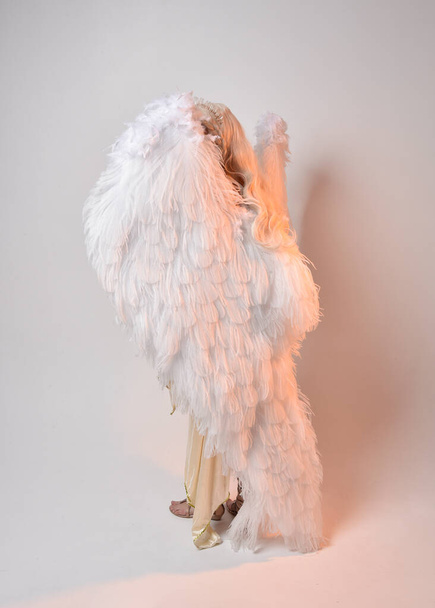 Retrato de larga duración de una hermosa mujer rubia con un disfraz de toga de diosa de fantasía con alas de ángel emplumadas. Saltando posan como volando, aislados sobre fondo blanco del estudio. - Foto, Imagen