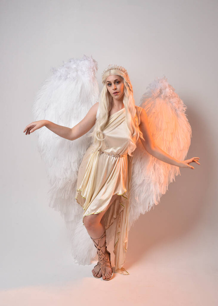 Tüylü melek kanatları olan fantezi tanrıça toga kostümü giyen güzel sarışın kadının tam boy portresi. Atlama pozu, beyaz stüdyo arka planında izole edilmiş.. - Fotoğraf, Görsel