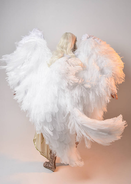 羽の天使の翼を持つファンタジーの女神トガ衣装を身に着けている美しいブロンドの女性の完全な長さの肖像画。飛行のようなジャンプポーズ、白いスタジオの背景に隔離された. - 写真・画像