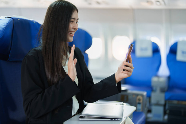 Mujer de negocios asiática pasajera sentada en avión de lujo de clase ejecutiva mientras trabaja usando teléfono inteligente móvil hablando o videoconferencia y concepto de viaje. - Foto, imagen