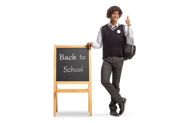 Full length portrait of a male african american student in a uniform leaning on blackboard with text back to school isolated on white background - Photo, image