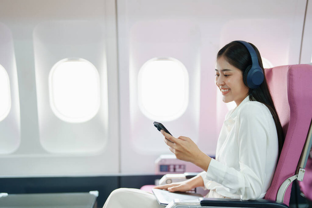 Joven asiática atractiva mujer de negocios pasajera sentada en un avión de lujo de clase ejecutiva mientras usa auriculares inalámbricos y usa un teléfono inteligente móvil para relajarse durante el vuelo. - Foto, Imagen