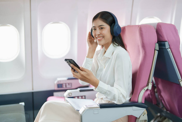 Młoda Azjatka atrakcyjny biznes kobieta pasażer siedzi na biznes klasy luksusowy samolot podczas noszenia słuchawki bezprzewodowej i za pomocą smartfona komórkowego na relaks podczas lotu. - Zdjęcie, obraz