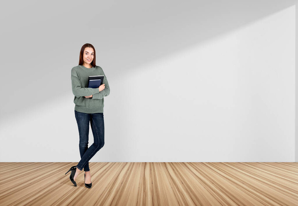 Attractive businesswoman wearing casual wear is standing holding notebook near concrete empty wall in background. Oak wooden floor. Concept of working process, taking notes for daily schedule - Photo, image
