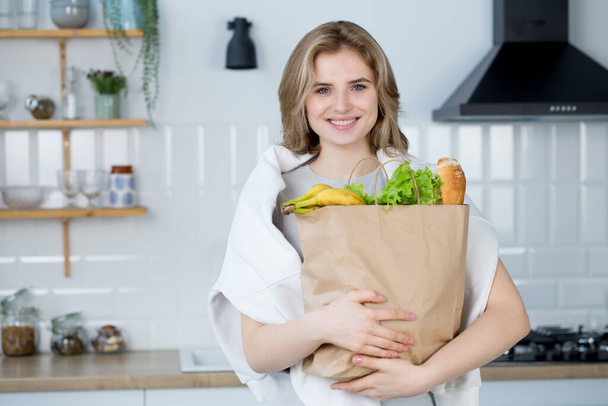 Güzel, genç, gülümseyen beyaz kadın modern mutfakta taze sebze ve baget dolu eko alışveriş çantası tutuyor.. - Fotoğraf, Görsel