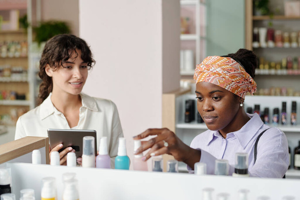 Joven consumidor femenino afroamericano eligiendo el producto beautycare de gran variedad mientras está de pie frente a la pantalla - Foto, imagen