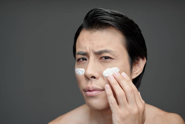 Frowning man applying moisturizer or sunscreen on face - Foto, imagen