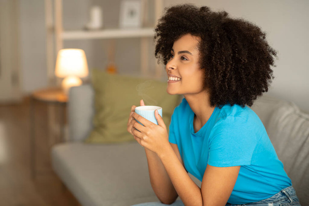 Froh millennial afrikanisch lockige Dame genießen Entspannung und Tasse Tee auf dem Sofa im gemütlichen Wohnzimmer Interieur, Blick auf Kopierraum. Guten Morgen, entspannen Sie sich abends allein, Kaffeepause und Stille - Foto, Bild