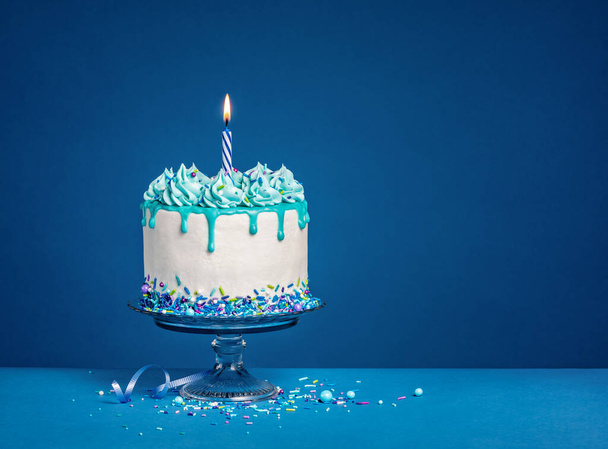 Λευκό κέικ γενεθλίων στάζει με teal ganache, ψεκάζει, και ένα αναμμένο κερί πάνω από ένα σκούρο μπλε φόντο. Αντιγραφή χώρου. - Φωτογραφία, εικόνα