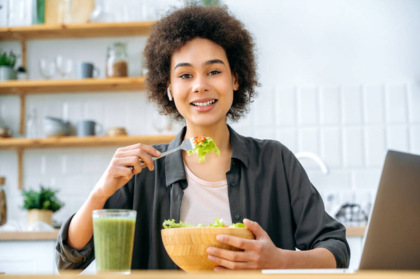 Mooi gezond Afrikaans Amerikaans meisje met kort krullend haar, zorgt voor haar gezondheid, eet gezond eten, zit in de keuken aan tafel, eet verse groentesalade, kijkt naar de camera, glimlacht - Foto, afbeelding