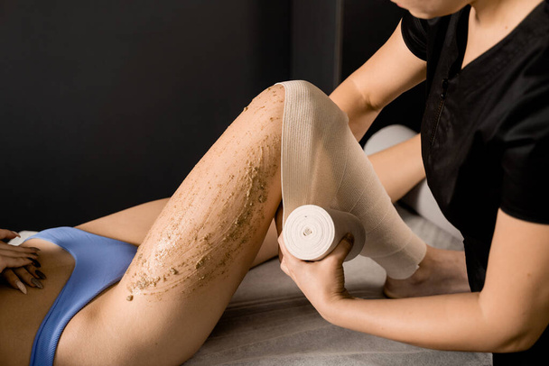 Bandage wrapping proces na het aanbrengen van scrub op benen van meisje voor styx wrapping en banding anti-cellulitis procedure in spa. Masseur doet scrub massage voor huidreiniging in spa - Foto, afbeelding