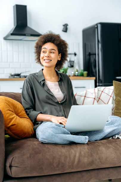 Vertical photo of a lovely young african american woman, student, freelancer, programmer, sit on a sofa in living room with a laptop, working online, typing on keyboard, looks away, dreaming, thinking - Φωτογραφία, εικόνα