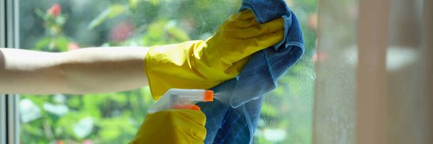 Woman hand in yellow gloves washes window with blue rag and detergent. Spring cleaning housework concept - Фото, изображение